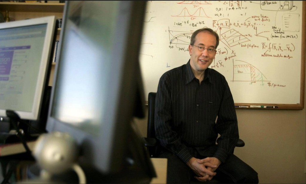 Andrew, smiling, seated in front of a whiteboard covered in equations, with computer monitors in the foreground.
